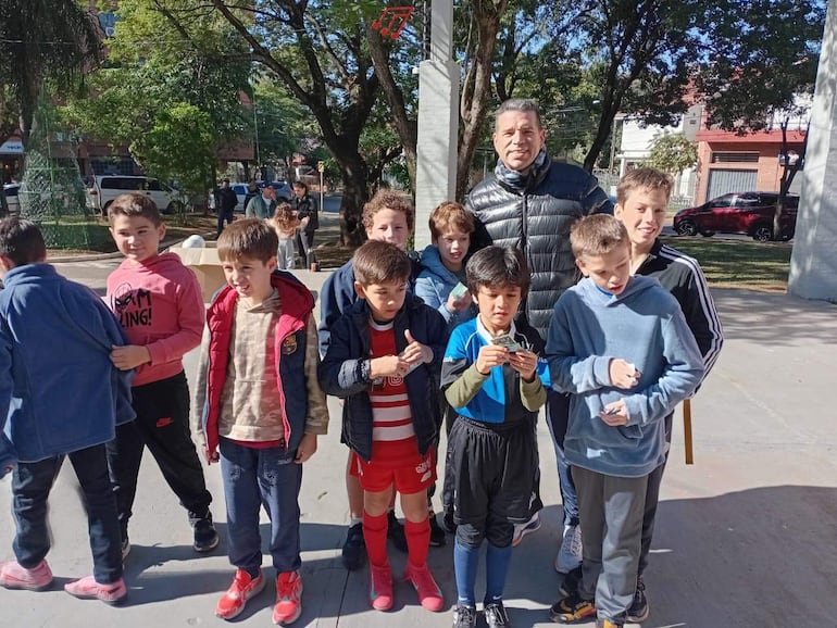 Diez niños y un adulto en la plaza, algunos sonríen, hay árboles y vehículos de fondo. Uno viste camiseta roja y blanca.