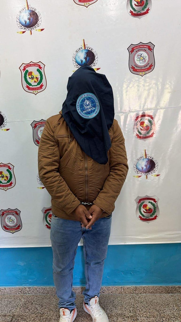 Hombre esposado de pie con chaqueta marrón y camiseta oscura en un ambiente policial con escudos visibles.