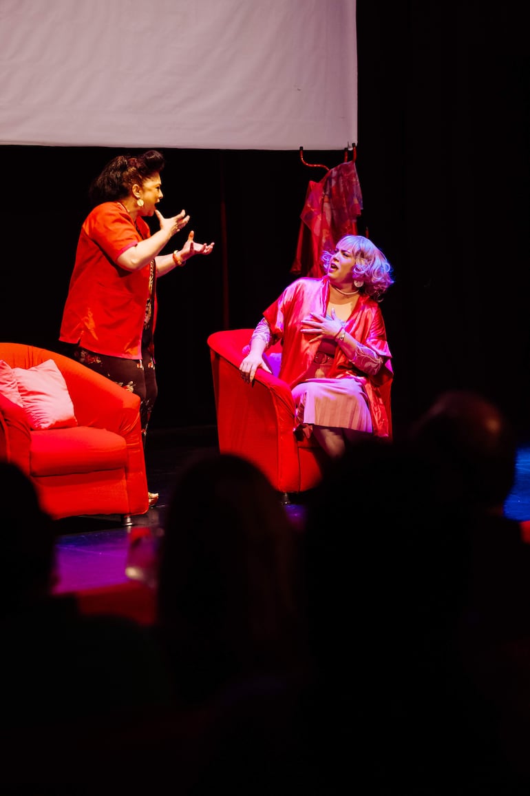 Actriz de cabello oscuro en camisa roja dialoga enérgicamente, mientras otra mujer de cabello rubio, en bata rosa, observa preocupada en un sillón.