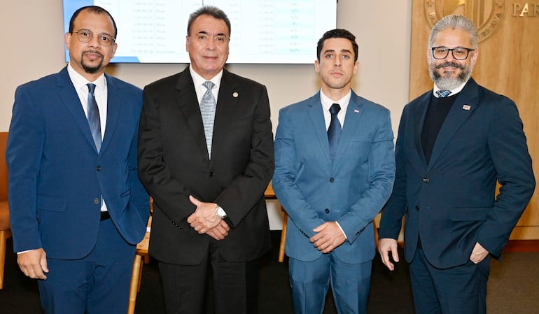Cuatro hombres en trajes formales discuten en sala de conferencias, con atención centrada en el orador en el centro.
