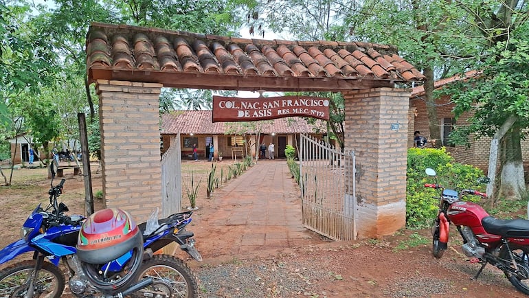 El colegio con entrada de ladrillo y tejado, con motocicletas estacionadas, en un entorno rodeado de árboles.