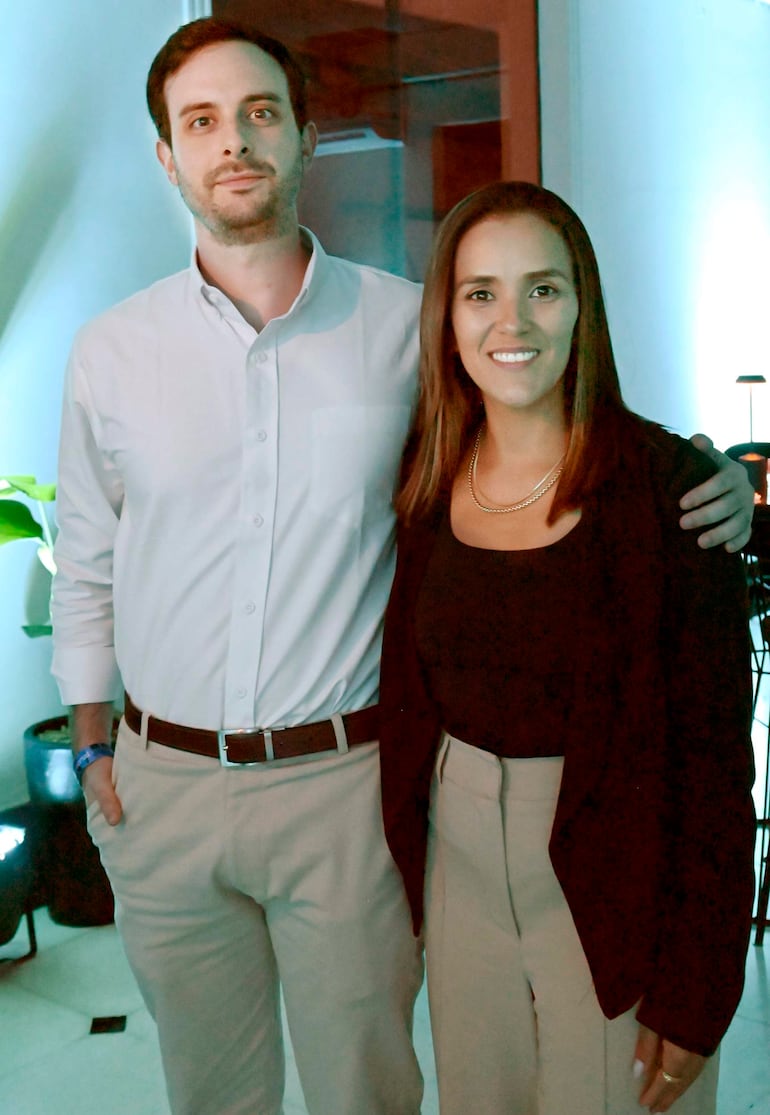 Hombre y mujer sonrientes en un entorno moderno, el hombre viste camisa blanca y pantalones claros, la mujer lleva blusa negra.