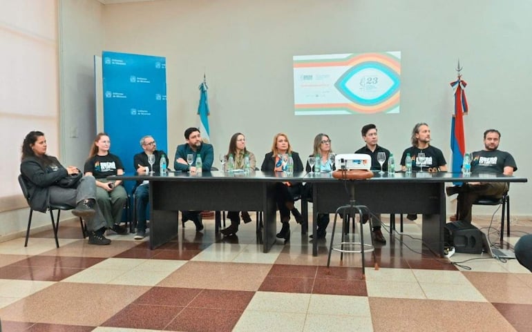 Nueve organizadores del festival sentados en mesa larga, vestidos de negro, atentos a una presentación, con bandera de Paraguay de fondo.