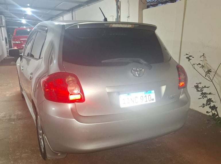 Toyota color plata estacionado en garaje, con faros traseros iluminados y vehículo rojo al fondo.