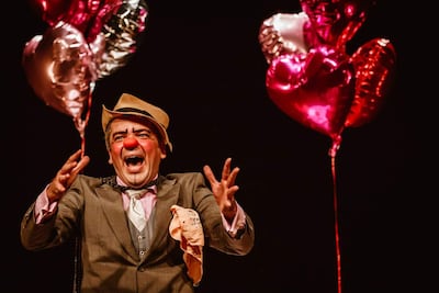Payaso en escenario oscuro, con maquillaje brillante y globos en forma de corazón, gesticulando con alegría.