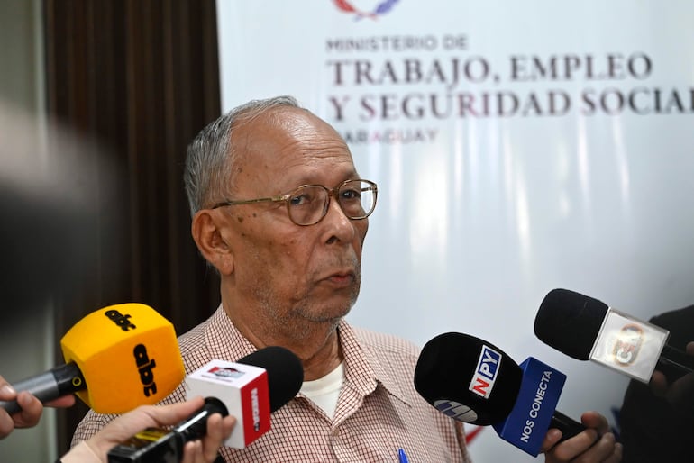Hombre mayor con gafas y camisa a cuadros, habla frente a micrófonos en una sala del Ministerio de Trabajo.