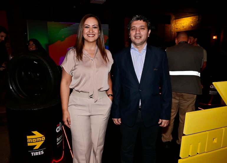 Mujer con blusa clara y pantalones beiges sonríe junto a hombre con chaqueta oscura, en ambiente festivo con iluminación colorida.