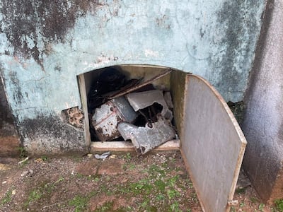 Abertura en pared azul deslavada muestra objetos metálicos oxidados y escombros en un piso de tierra y hierba.