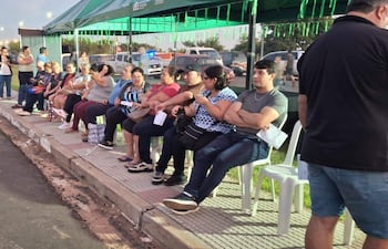 Grupo de personas sentadas, algunas conversando y otras en sus teléfonos, en un ambiente urbano con vehículos de fondo.