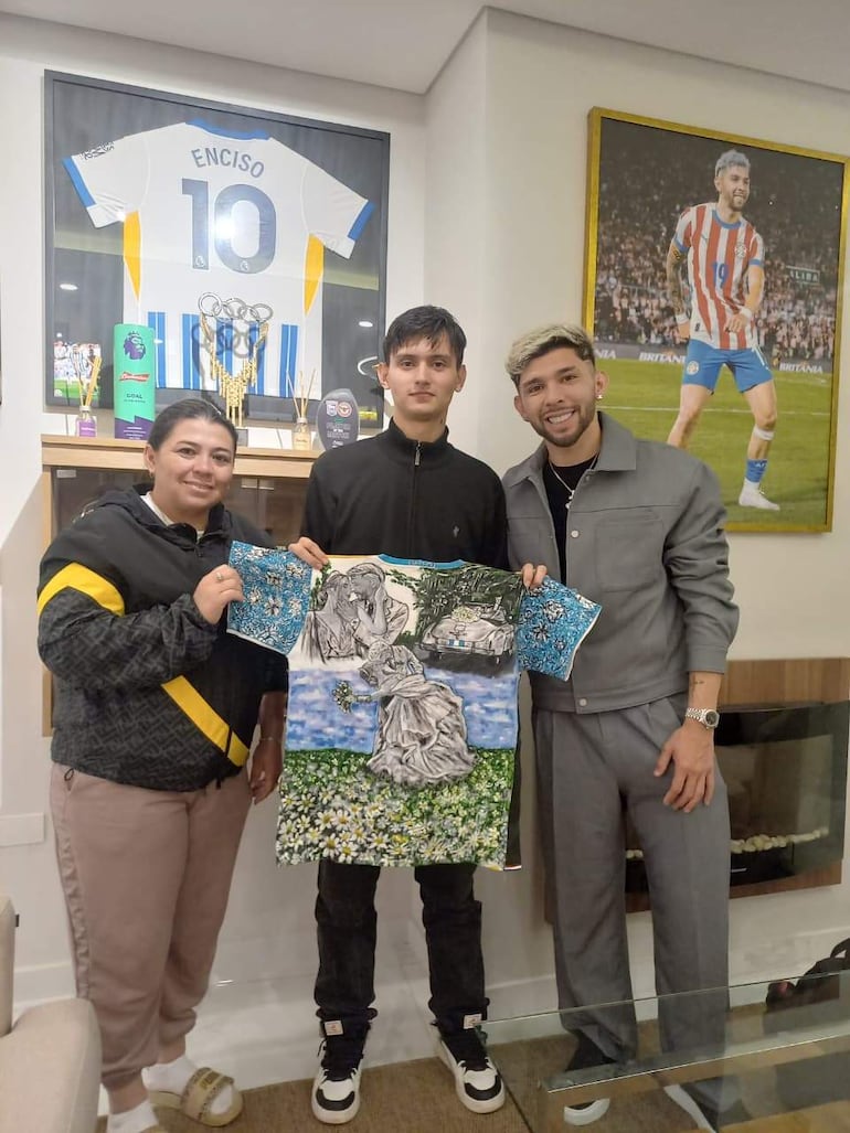 Ignacio Villalba sonriente, sosteniendo una camiseta con el nombre Enciso, acompañado de Julio Enciso y su madre Angelina Espínola en un ambiente acogedor.