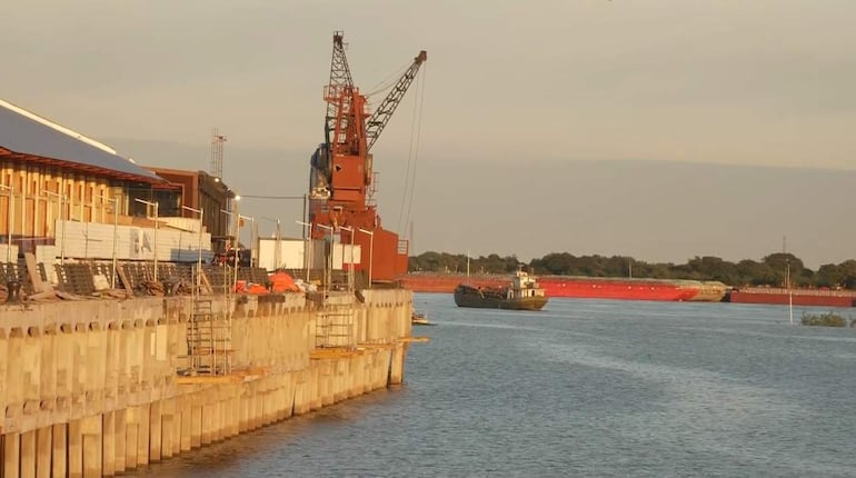 Entorno portuario con grúa roja, muelles industriales y barco en el río Paraguay rodeado de árboles.
