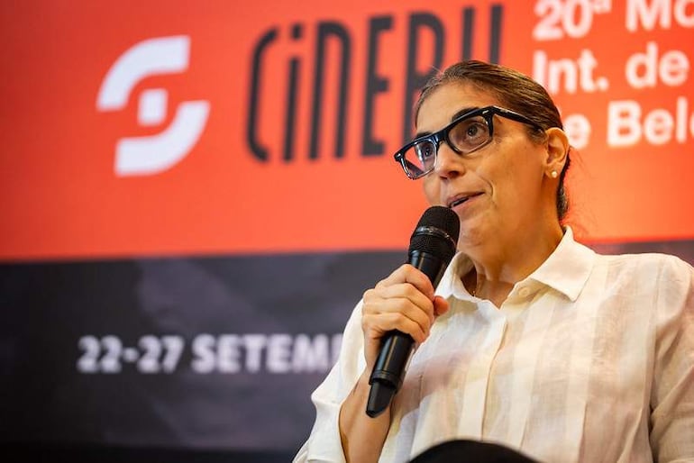 Paz Encina, con vestimenta clara, sostiene un micrófono en un salón decorado en rojo y negro, mientras se lee 'CINERU' en el fondo.