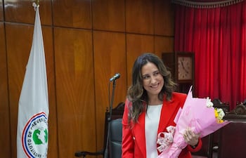 Bettina Albertini, sonriente con un blazer rojo y ramo de flores, se encuentra en un ambiente de oficina con documentos y una bandera institucional.