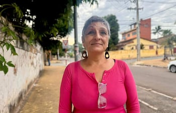 Mujer de cabello corto y canoso con camisa rosa, de pie en la vereda, mirando a la cámara con edificios coloridos de fondo.