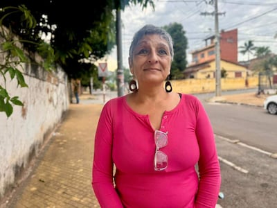 Mujer de cabello corto y canoso con camisa rosa, de pie en la vereda, mirando a la cámara con edificios coloridos de fondo.