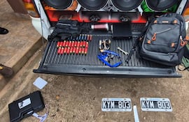 Maletero de auto con cartuchos rojos, destornillador, computadora y mochila sobre superficie de tierra.