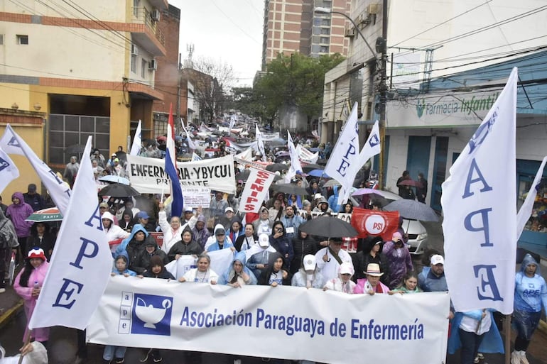 Manifestantes con paraguas y pancartas en apoyo a la Asociación Paraguaya de Enfermería, en un ambiente lluvioso.