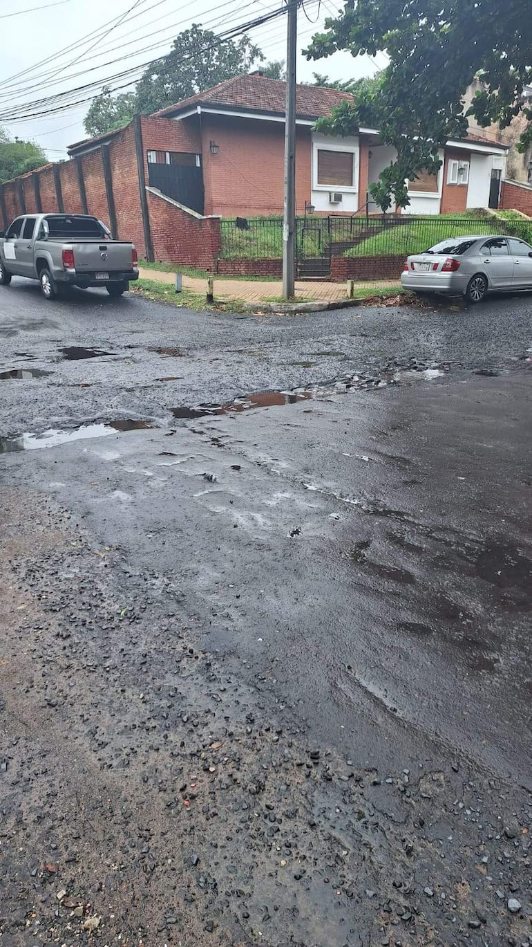 Calle en mal estado con vehículos estacionados, pavimento dañado y agua acumulada, rodeada de casas de ladrillo y postes de luz.