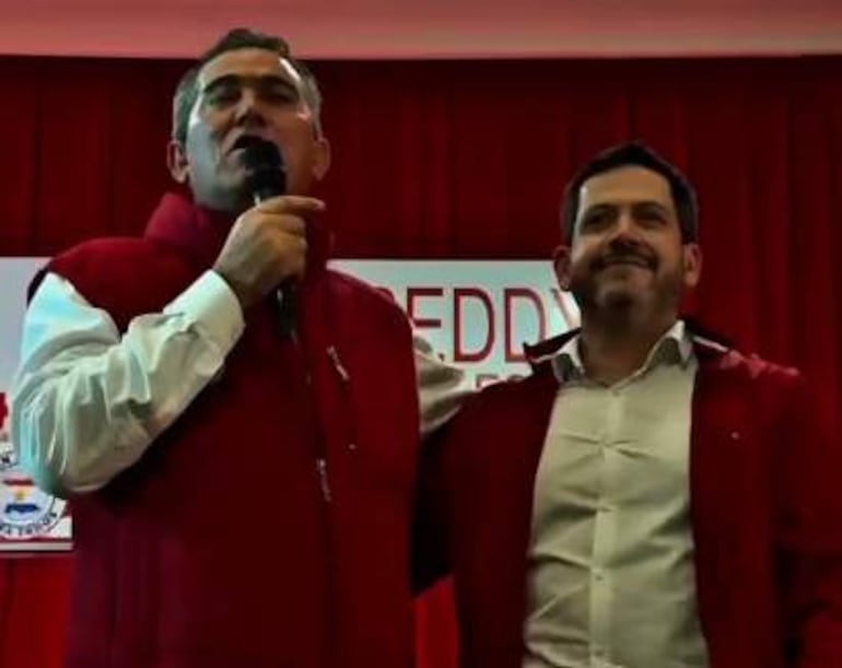 Raúl Latorre sonríe mientras sostiene un micrófono, junto a Leonardo Saiz en abrigos rojos, en un entorno decorado con tela roja.
