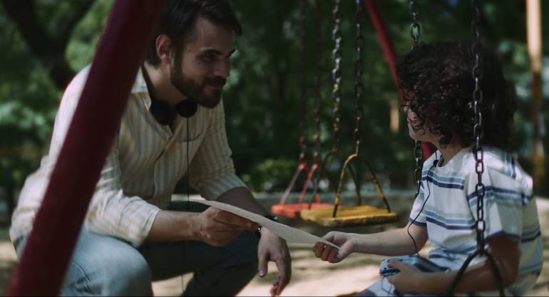Adulto con barba y camisa de rayas amarillas se inclina hacia un niño con camiseta de rayas azules en un columpio.