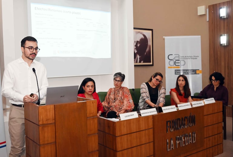 Presentador en camisa blanca frente a proyección, mujeres sentadas con atenta expresión y colores variados en su vestimenta.