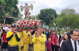 Dos hombres en uniformes amarillos cargan un pedestal con la figura de San Miguel, mientras una mujer sostiene un cartel con la Virgen del Carmen.