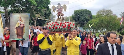 Dos hombres en uniformes amarillos cargan un pedestal con la figura de San Miguel, mientras una mujer sostiene un cartel con la Virgen del Carmen.