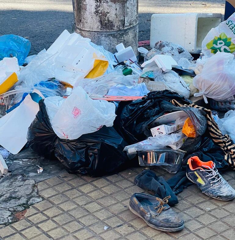 Montículo de basura en una acera con bolsas plásticas y zapatos sucios visibles, ambiente urbano descuidado.