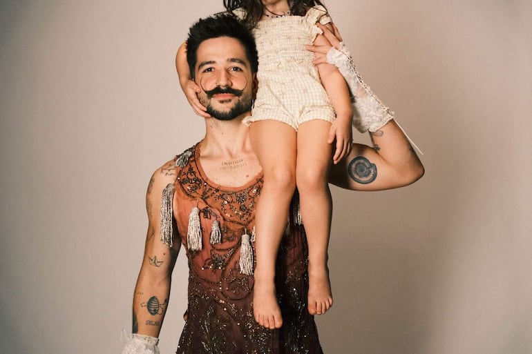Camilo con barba y atuendo brillante sonríe mientras sostiene a su hija, que lleva un mono claro sobre sus hombros, en un fondo neutro.