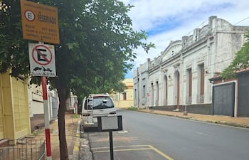 Calle tranquila con señal de estacionamiento reservado; cielo nublado y edificio clásico al fondo.