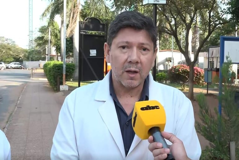 Diego Gamarra, con bata blanca y micrófono de ABC Color, habla frente a una cámara entre árboles en un ambiente urbano.