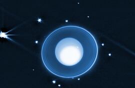 En el centro está el planeta Urano, un globo azul con una gran mancha blanca que ocupa la mayor parte del planeta. Está rodeado por una serie de anillos que se vuelven más brillantes hacia el exterior. Más allá de los anillos se distinguen 14 lunas que aparecen como puntos de luz. Las lunas varían en brillo, siendo la más brillante, Miranda, visible justo encima de la posición de las 9 en punto.