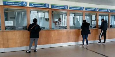 Tres personas de espaldas en una farmacia: dos mujeres con abrigos negros y un hombre con chaqueta oscura, en ambiente de atención médica.