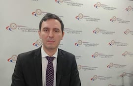 Leandro Villalba sentado en una mesa, vistiendo traje oscuro y corbata a rayas moradas, frente al logo de la Contraloría General de la República.
