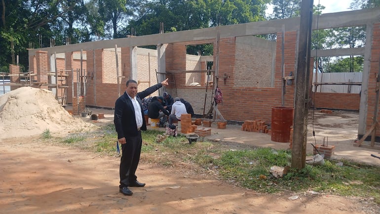 Jorge Servín en traje oscuro señala hacia la obra situada detrás, con trabajadores en la construcción y ladrillos apilados en el fondo.