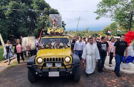 Un jeep amarillo decorado avanza en la procesión, seguido por feligreses y un sacerdote con sotana blanca en un ambiente comunitario.