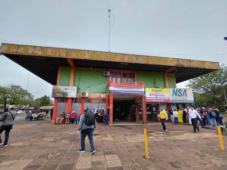 Hombre con chaqueta gris y bolso negro observa la terminal, mientras un grupo de personas espera en fila bajo un cielo nublado.