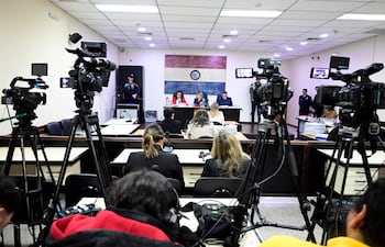 Tres personas en la mesa: dos mujeres, una de blanco y otra de rojo, y un hombre en traje. Oficiales de seguridad al fondo.