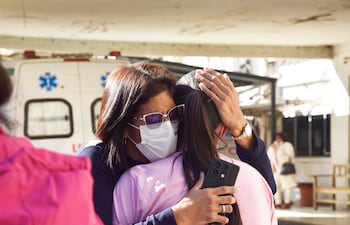 Dos mujeres, una con blusa rosa y otra con mascarilla, se abrazan mientras una ambulancia se ve al fondo.