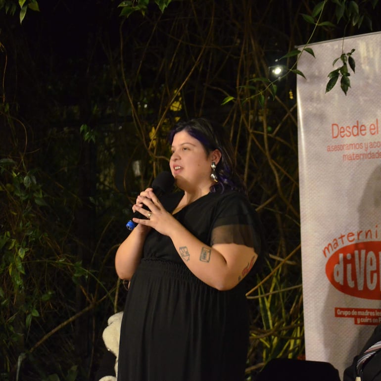 Mujer de cabello corto con puntas púrpuras, vistiendo un vestido negro, sostiene un micrófono mientras habla en un ambiente iluminado con plantas.