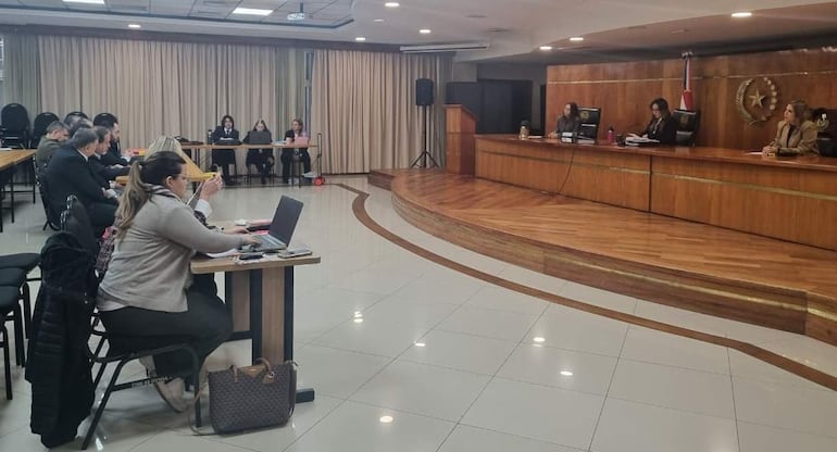 Mujer con laptop al frente en sala de audiencias, rodeada de personas atentas y juezas en el fondo con documentos y agua sobre la mesa.