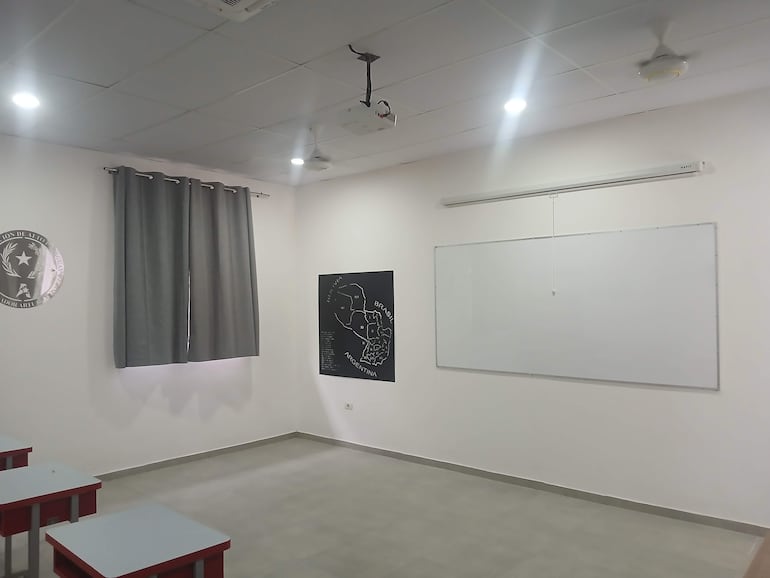 Aula vacía con pizarra blanca, mural de mapa y cortina gris, preparada para clases en ambiente luminoso.