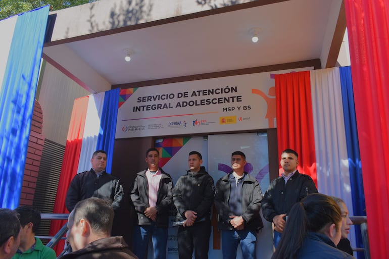 Cinco hombres de pie en el exterior, vestidos con ropa oscura, ante un cartel del Servicio de Atención Integral Adolescente, con decoración en rojo, blanco y azul.