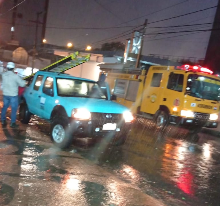 Persona con sombrero blanco manipula objeto en camioneta azul, mientras se observa vehículo amarillo de bomberos bajo la lluvia.