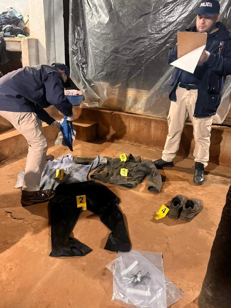 Agentes de policía en uniforme manipulan prendas como evidencia en un entorno de investigación con paredes cubiertas de plástico.