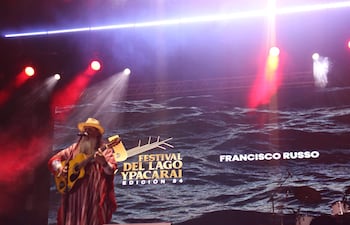 Francisco Russo en escenario, tocando guitarra y usando un sombrero amarillo. Luces de colores iluminan el ambiente festivo.
