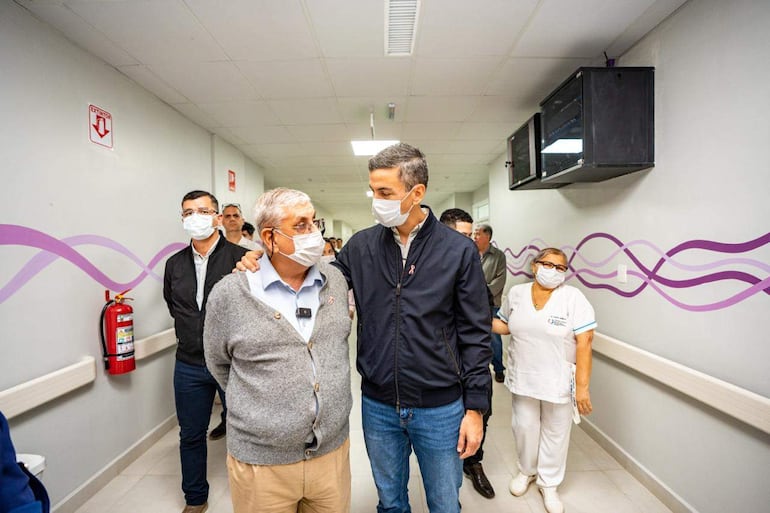 Peña con chaqueta negra dialoga con un hombre canoso en un hospital, rodeados por personal de salud y otros observadores.