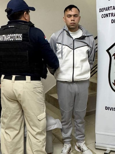 Hombre de pie en custodia, con ropa deportiva gris, mientras otro en vestimenta negra le habla en ambiente sobrio.