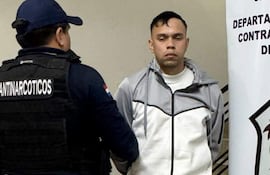 Hombre de pie en custodia, con ropa deportiva gris, mientras otro en vestimenta negra le habla en ambiente sobrio.