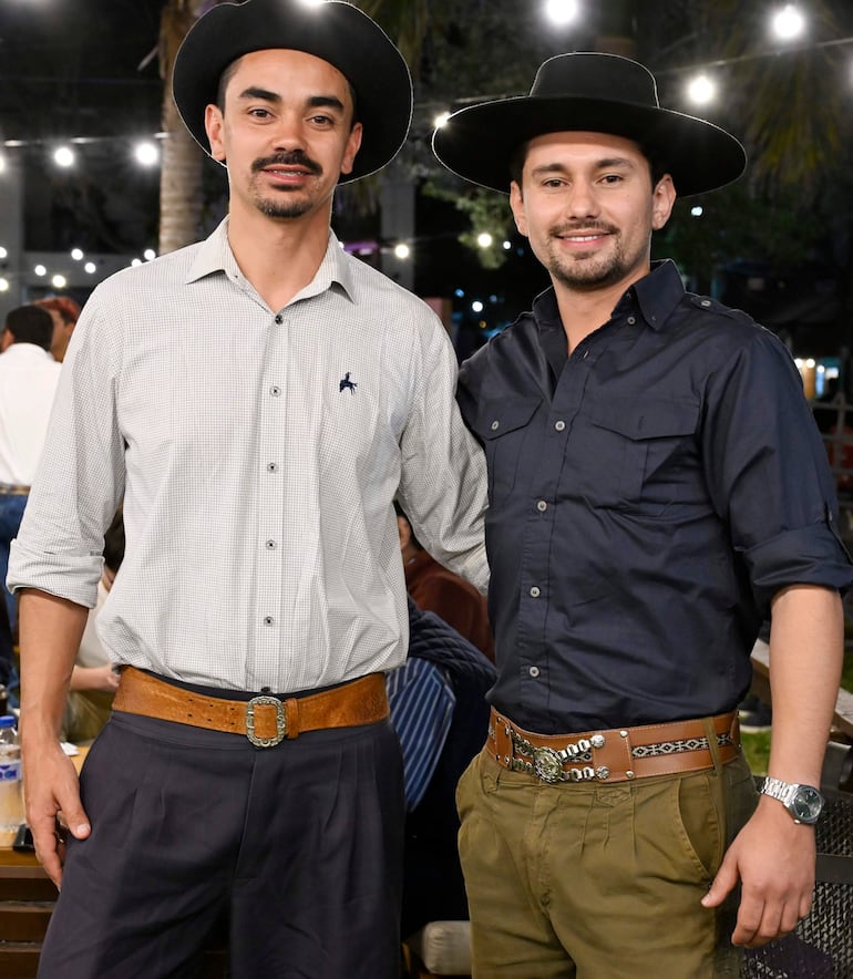 Dos hombres sonríen en un ambiente cálido, uno con camisa de cuadros y otro con camisa oscura, ambos con sombreros de ala ancha.